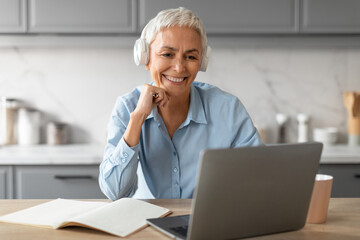 Cheerful senior lady in headphones watching lecture on laptop indoor