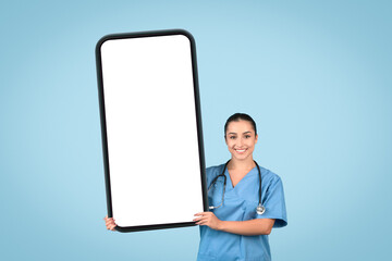 Cheerful young female nurse with an oversized cellphone displaying white blank screen for medical adverts and offers