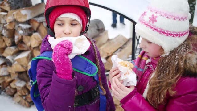 Two girls friends eat pasties near firewood outdoor at winter