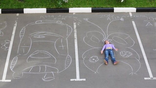 Girl lies on big butterfly drawn by chalk near draw car on asphalt
