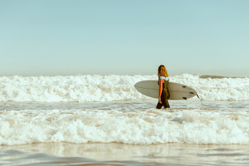 Side view of female surfer in wetsuit with his surfboard entering the sea. Surfing on waves