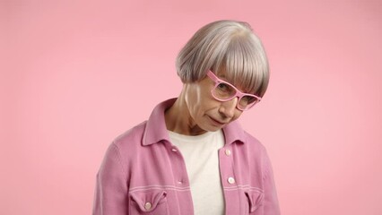 A woman of advanced age appears embarrassed, her cheeks matching the blush of her pink outfit, which seamlessly blends into the pink background. Camera 8K RAW. 