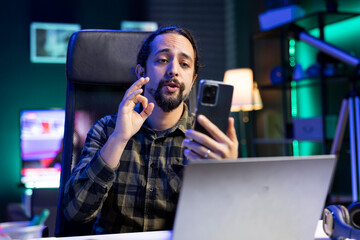 Bearded man sitting in front of laptop while having a virtual conversation through his smartphone. Male freelancer using his cell phone for video call, to assure his customers.