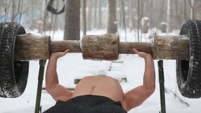 Bare-chested guy lies at lounger and pushes up wooden barbell