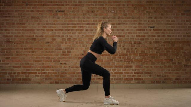 Split squat. Fitness trainer in black sportswear demonstrates the correct execution of the exercise, split squat. She is in an empty hall with a brick wall. Camera 8K RAW.