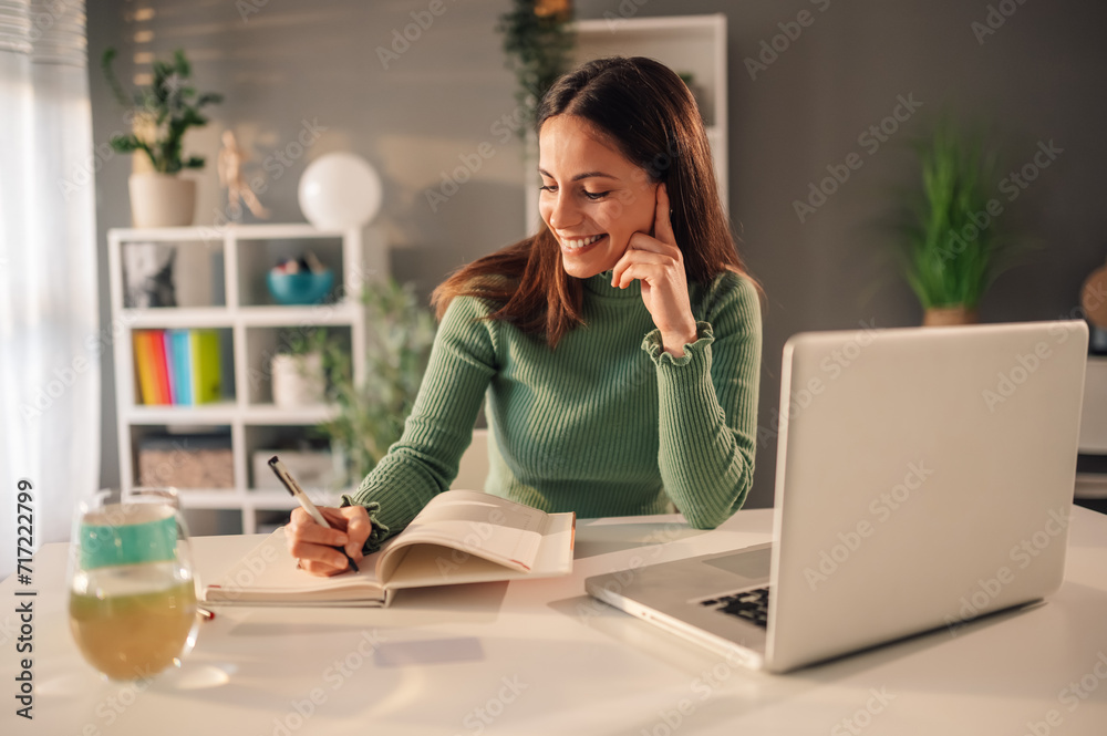 Wall mural smiling young woman working at home with laptop and papers on a desk