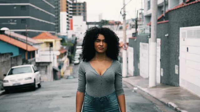 One confident young black woman with curly hair standing outside in urban setting looking at camera with stern solemn expression. South American, 20s person in street portrait face