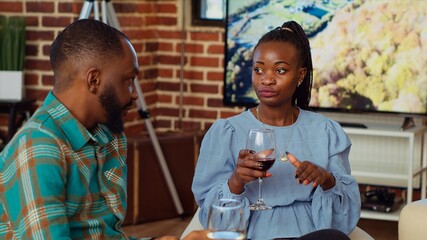 African american man at social gathering talking with his girlfriend on couch while enjoying glass of wine. Husband and wife guests at apartment party having pleasant conversation in brick wall home