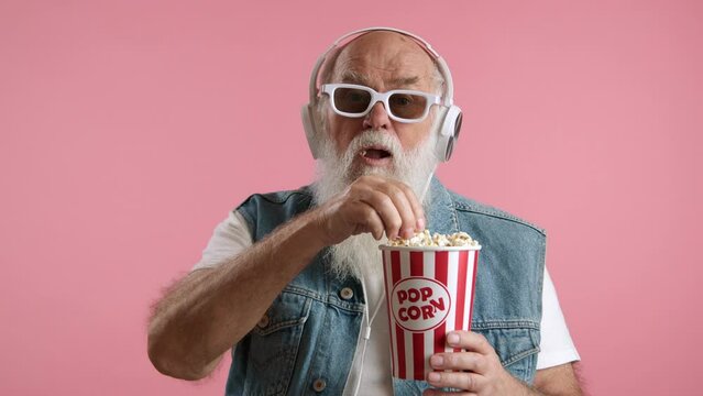 A pensioner in youthful attire, wearing a white t-shirt and denim vest, with headphones, 3D glasses, and popcorn, is captivated by a movie in a cinema. Pink background. Camera 8K RAW. 