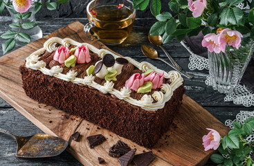 Delicious biscuit cake "Fairy Tale" decorated with dessert mushrooms and flowers with chocolate cream on dark background with cup of tea and spring flowers. Sweets, dessert and pastry, top view