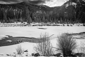 Classic black and white landscape art of the partially frozen north fork Shoshone River and snow-covered mountain landscape with storm clouds at sunset in Wyoming, USA. 