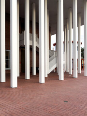 The exterior entrance to a building with a white metal staircase to a second floor. In front of the stairs are multiple vertical white smooth pillars and columns. The ground is covered in red brick.