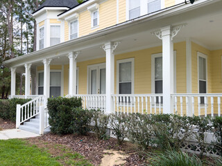 The facade of a large yellow and white colored wooden middle class house with a wraparound porch on...