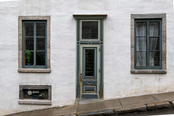 The exterior of a vintage white masonry wall with multiple glass windows, half glass single door, a transom window over the entrance and green colored trim. The facade of the house is on a steep hill.