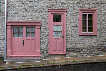 The exterior wall of a grey colored stone building with two closed glass windows and a tall vintage pink-colored wooden door. The wall is crumbling. There's a white curtain on the door's glass window.