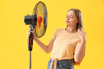 Sweaty young woman with electric fan on yellow background