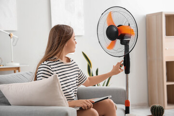 Young woman with laptop and blowing electric fan at home