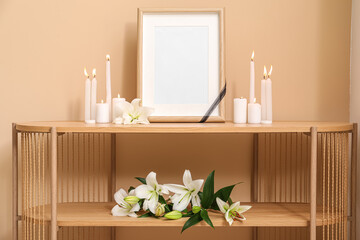 Blank mortuary frame, burning candles and white lily flowers on wooden table near color wall