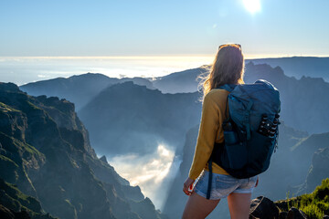 Female backpacker tourist  overlooks a deep, cloud-covered valley and enjoys the breathtaking view of the volcanic mountain landscape. Pico do Arieiro, Madeira Island, Portugal, Europe. - obrazy, fototapety, plakaty