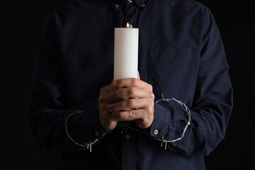 Young Jewish man with barbed wire and burning candle on black background, closeup. International...