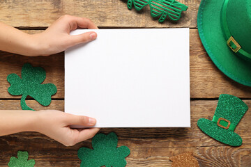Female hands with blank card, leprechaun hat and paper clovers on wooden background. St. Patrick's...