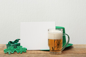 Blank card with mug of beer, gift box and plastic eyeglasses on wooden table against white background. St. Patrick's Day celebration