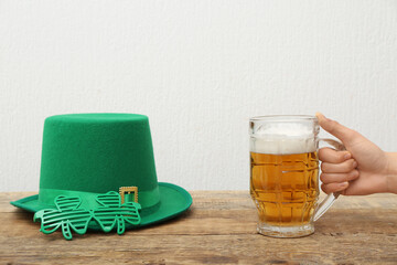 Female hand with mug of beer, leprechaun hat and plastic eyeglasses on wooden table against white...