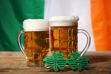 Mugs of beer and plastic eyeglasses on wooden table against flag of Ireland. St. Patrick's Day celebration