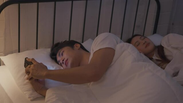 Asian man secretly chatting to mistress while lying in bed with girlfriend. 