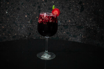 Imagen horizontal de una copa de vino o clericot con frutos rojos adornada con una fresa partida a la mitad sobre una mesa y entorno obscuros