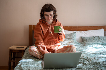 Teen girl watching series movie on laptop sitting in bed drinking tea at home. Woman introvert in...