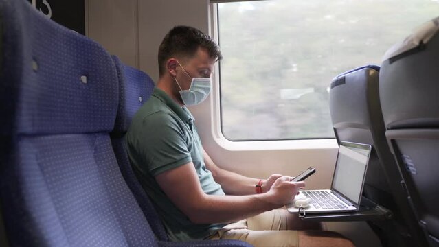 Man texting on phone holding laptop, commuter train in protective mask during coronavirus pandemic. Theme travel by rail, new rules life during COVID 19. Male uses smartphone and laptop in train.