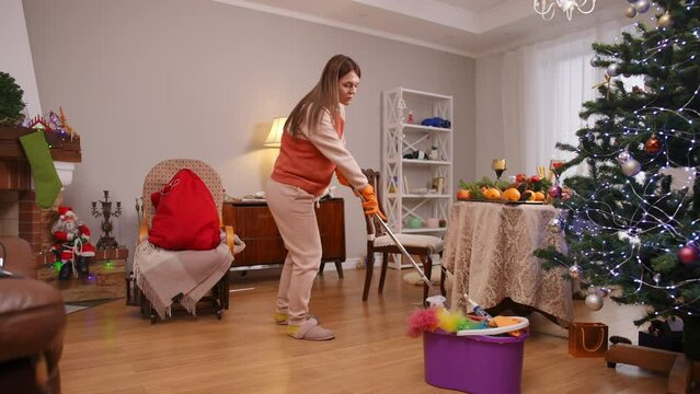 A pregnant woman wipes water with a mop from a wooden floor in the living room before the Christmas holidays. The woman is in a good mood and smiling