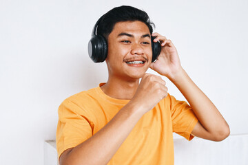 Excited young Asian man with braces teeth, listening music from headphones and sing