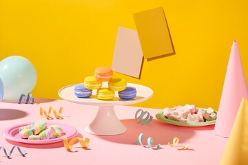 Macaron on a white plate, marshmallow on a round plate, ribbons, balloons and birthday hats decorated around. The note pads are stuck on a yellow background.