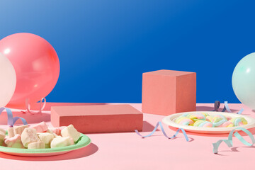 Two empty podiums for display. Balloons, ribbons and two marshmallow dishes on a pink surface with a blue background. Copy space with front view.