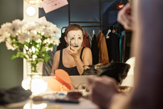 Mirror reflection portrait of female mime performer doing makeup backstage preparing for show in theater, copy space
