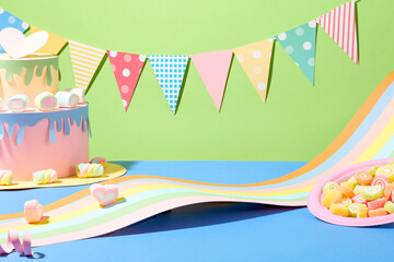 A string of birthday decorations on a green background. A birthday cake made from colored paper, a strip of colorful paper and a plate of gummy candy on a blue surface. Front view.