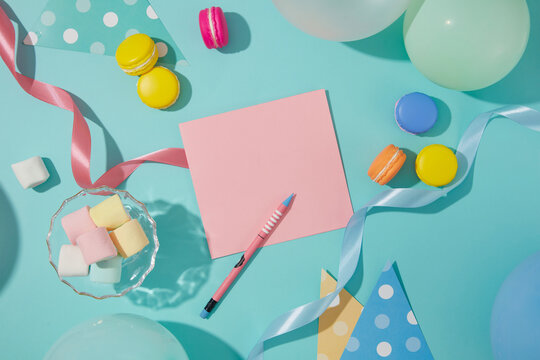 A pencil sits next to a pink card. Marshmallows, ribbons, balloons and macarons are decorated around the blue background. Free space to design birthday wishes.