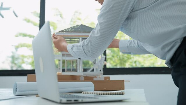 Close up of professional male architect engineer measure house model and using laptop analysis data at meeting table with house model and map . scatter around. Focus on hand. Immaculate.
