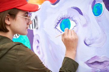 Female painter in red cap draws surreal picture with paintbrush on canvas for outdoor street exhibition, close up side view of female artist apply brushstrokes to canvas, symphony of art creativity