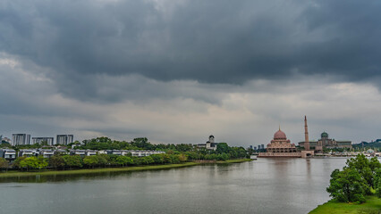 A calm urban lake. Green vegetation on the banks. A pink  Putra Mosque is visible in the distance. Minaret and domes against a cloudy sky. Malaysia. Putrajaya.