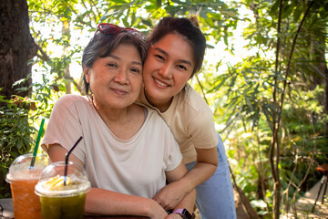 Asian Thai Chinese daughter cuddling and hugging her elder mother, both woman looking at camera, drinking beverages at cafe garden, spending time together.
