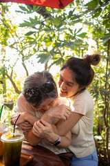 Asian Thai woman daughter hugging her elder mother, playful and laughing together while sitting at cafe garden.