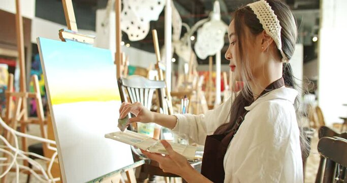 Beautiful asian stylish female artist painting picture in art studio. Young woman painter using a sponge applying paint on canvas creating artwork sunset landscape. imagination and creativity.