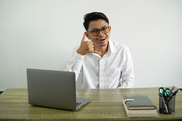 young asian businessman in a workplace making phone gesture with fingers. call me back sign, wear...