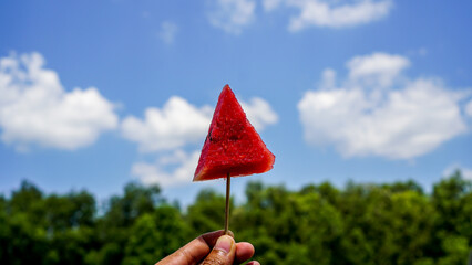Female Hand holding piece of fruit picks watermelon lift up in the sky on a bright day with blue...