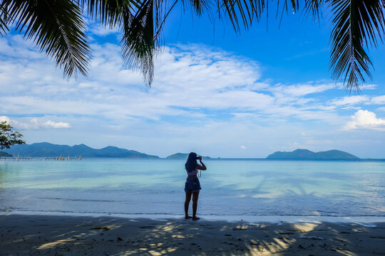 A side view of a young asian woman using her smartphone up and pointing it to take pictures view of lake shore with sea in background. Happy woman taking photos to the sea with smartphone camera