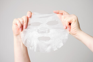Female hands hold fabric sheet mask on gray background. - obrazy, fototapety, plakaty