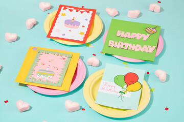 Four greeting cards with four different designs are placed on four round paper plates. Heart-shaped marshmallow and confetti decorated on a pastel blue background.